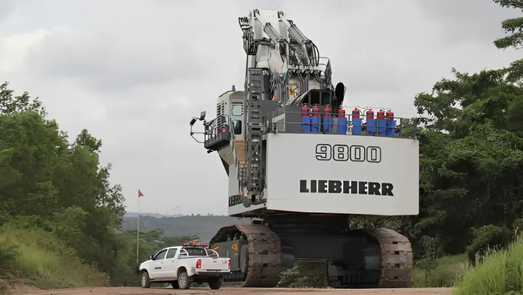 A side-profile graphic of the Liebherr R9800 sitting next to a standard pickup truck and a 3-story house to demonstrate its massive physical scale and track dimensions