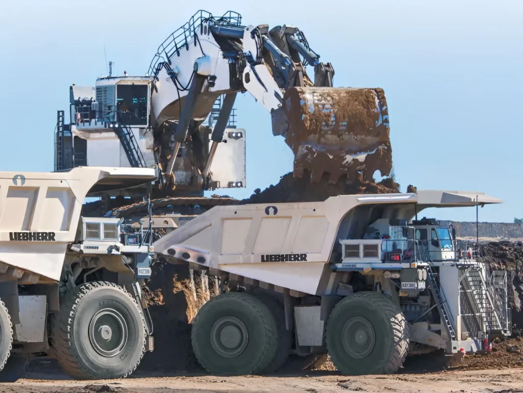 A wide-angle shot showing the Liebherr R9800 loading a 300-ton haul truck, while two other trucks wait nearby, illustrating the cost-per-ton logistics of a continuous mining cycle